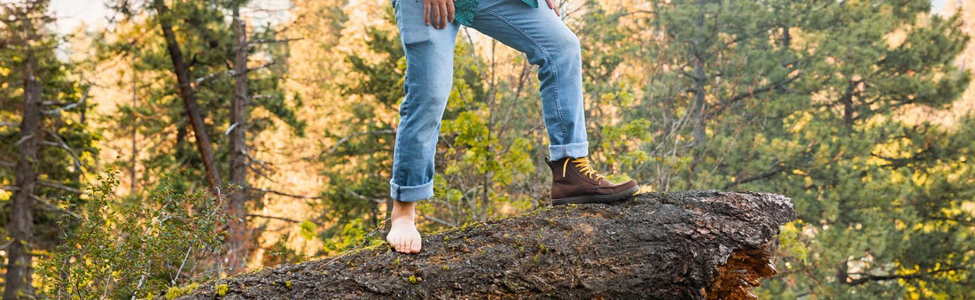 Persona de pie sobre un tronco en la naturaleza, con un zapato en un pie y el otro descalzo, destacando la versatilidad del calzado barefoot de BareMotion